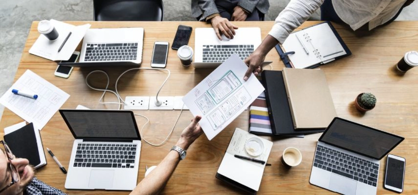 Photo en plongée sur un bureau de coworking, contenant 4 ordinateurs et 3 personnes autour de la table.